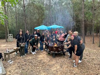 Group of about twenty people gathered around a metal fire pit in a pine forest campground, roasting marshmallows on long sticks under blue umbrellas and smiling during a cozy outdoor campfire gathering.