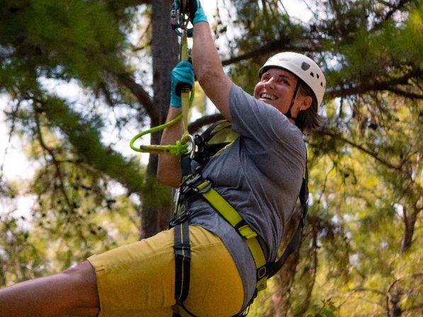 Smiling person in a helmet and neon safety harness suspended on a zip line in a sunlit pine forest — outdoor ropes-course adventure.