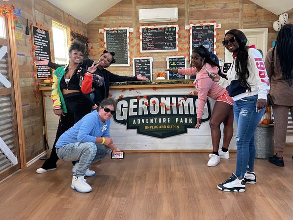 Group of friends posing cheerfully at an adventure-park ticket counter inside a rustic wooden cabin decorated with autumn leaves and chalkboard menus.