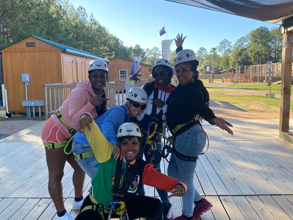 Five friends in helmets and safety harnesses pose and smile on a wooden zipline platform at an outdoor ropes course with cabins and pine trees on a sunny day.