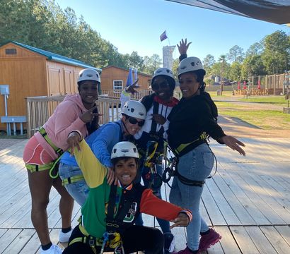 Five friends in helmets and safety harnesses pose and smile on a wooden zipline platform at an outdoor ropes course with cabins and pine trees on a sunny day.