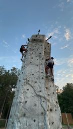 Two climbers scaling a tall outdoor artificial rock-climbing wall at dusk under a blue sky with scattered clouds and trees in the background