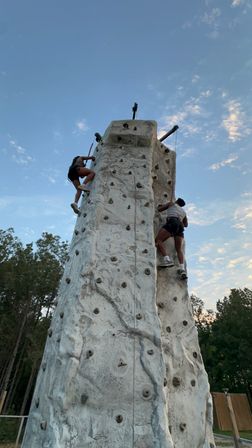 Two climbers scaling a tall outdoor artificial rock-climbing wall at dusk under a blue sky with scattered clouds and trees in the background