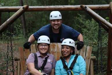 Three smiling adults wearing white helmets and safety harnesses sitting on a wooden swing at an outdoor adventure park in a forested area