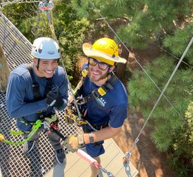 Two people wearing helmets and full-body harnesses, smiling and giving a thumbs-up on a high ropes course platform surrounded by pine trees, clipped into zipline cables with carabiners and safety lines.