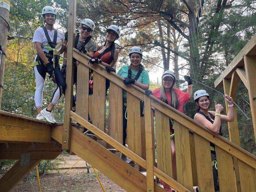 Six people in helmets and harnesses smile and give thumbs-up on a wooden stair platform at an outdoor zipline/ropes course in a forest setting.