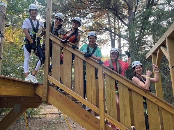 Six people in helmets and harnesses smile and give thumbs-up on a wooden stair platform at an outdoor zipline/ropes course in a forest setting.