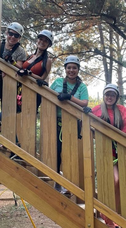 Six people in helmets and harnesses smile and give thumbs-up on a wooden stair platform at an outdoor zipline/ropes course in a forest setting.