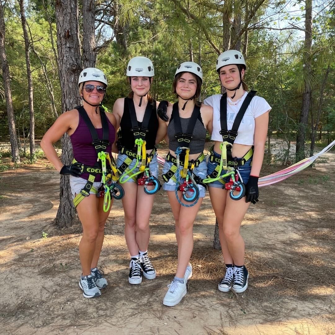 Four people in helmets and safety harnesses smiling and posing in a pine forest before a zipline on a sunny outdoor ropes-course adventure