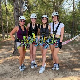 Four people in helmets and safety harnesses smiling and posing in a pine forest before a zipline on a sunny outdoor ropes-course adventure