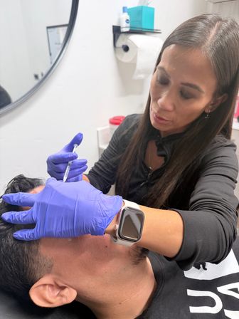 Close-up of a clinician wearing purple gloves administering a cosmetic facial injection to a patient’s forehead in a medical spa treatment room.
