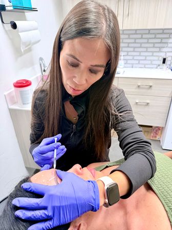 Close-up in an aesthetic clinic: clinician wearing purple gloves gently administers a facial injectable to a reclining patient’s forehead with visible guideline markings.