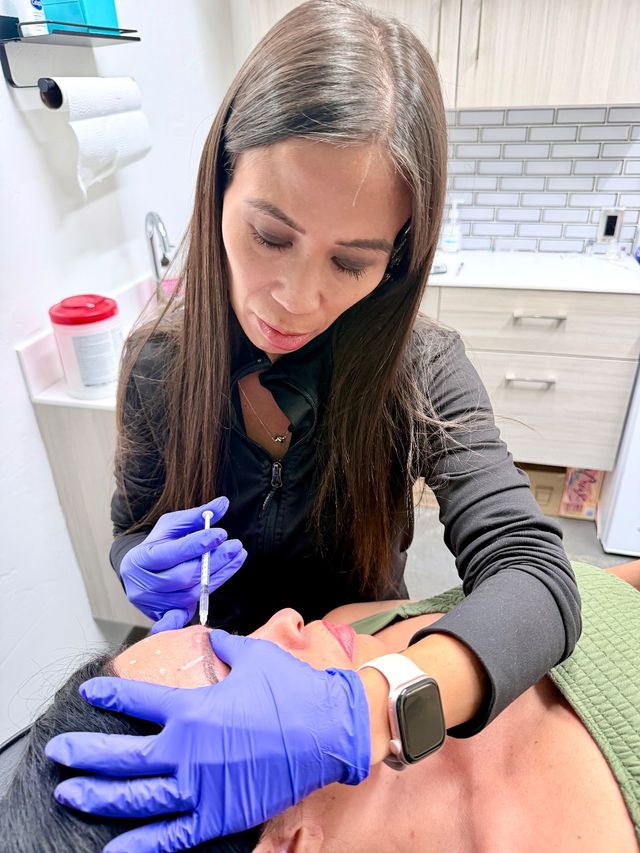 Close-up in an aesthetic clinic: clinician wearing purple gloves gently administers a facial injectable to a reclining patient’s forehead with visible guideline markings.