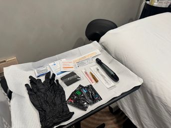Aesthetic clinic treatment tray with black nitrile gloves, sterile prep pads, alcohol wipes, surgical sponges, microneedling pen and injectable vials beside a white treatment bed.