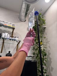 Gloved hands preparing an IV drip and syringe in a bright wellness clinic treatment room with skincare shelves and hanging greenery.
