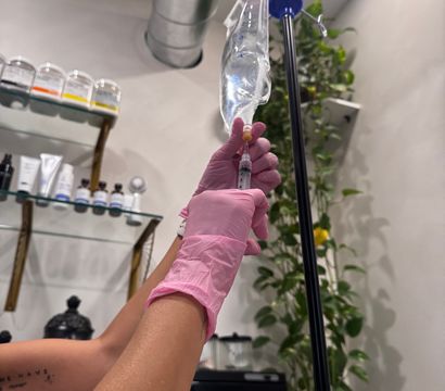 Gloved hands preparing an IV drip and syringe in a bright wellness clinic treatment room with skincare shelves and hanging greenery.