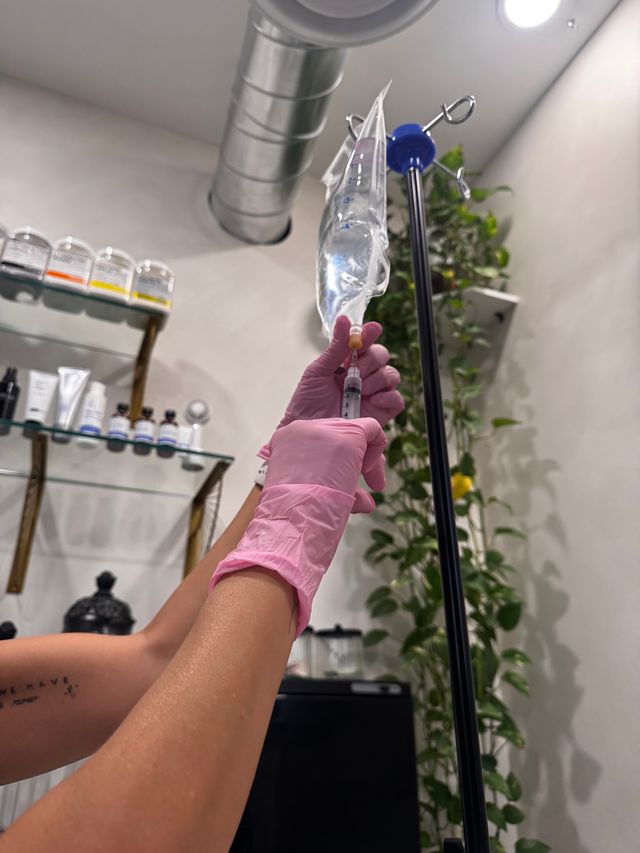 Gloved hands preparing an IV drip and syringe in a bright wellness clinic treatment room with skincare shelves and hanging greenery.