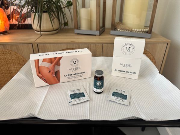At-home body peel kit staged on a disposable treatment drape — vial, two prep towelettes and boxed post-peel care on a spa table with wood decor and potted plant in the background.