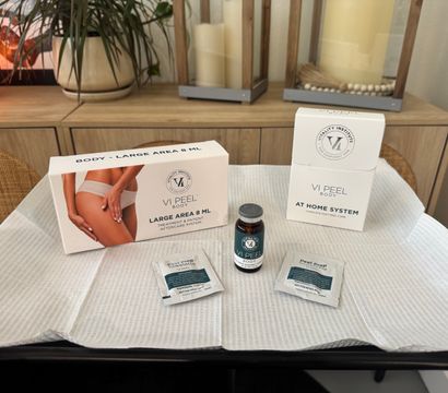 At-home body peel kit staged on a disposable treatment drape — vial, two prep towelettes and boxed post-peel care on a spa table with wood decor and potted plant in the background.