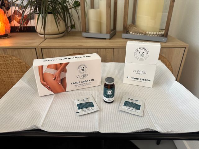 At-home body peel kit staged on a disposable treatment drape — vial, two prep towelettes and boxed post-peel care on a spa table with wood decor and potted plant in the background.