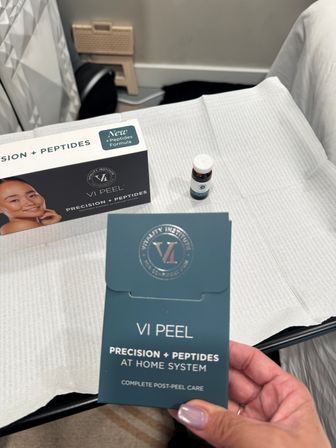 VI Peel Precision + Peptides at‑home system brochure and a small vial staged on a treatment table in a skincare clinic, hand with light pink manicure holding the brochure — post-peel care kit.