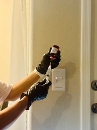 Close-up in a home interior: gloved hands draw medication from a small pink-capped vial into a syringe against a beige wall beside a light switch and door handle.