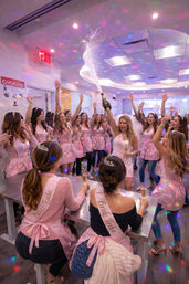 Bachelorette party in a bright indoor venue — bride in a veil pops champagne as a crowd of women in pink "Bride Tribe" sashes and tiaras cheer under colorful disco lights.