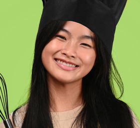 Smiling young cook wearing a black chef hat and holding a whisk against a bright green background