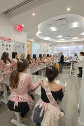 Bachelorette baking class in a bright modern kitchen studio, women wearing pink "Bride Tribe" sashes and tiaras seated at stainless worktables with dough and champagne while an instructor demonstrates.