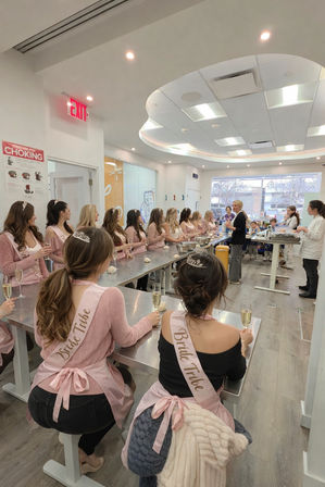 Bachelorette baking class in a bright modern kitchen studio, women wearing pink "Bride Tribe" sashes and tiaras seated at stainless worktables with dough and champagne while an instructor demonstrates.