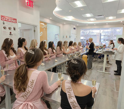 Bachelorette baking class in a bright modern kitchen studio, women wearing pink "Bride Tribe" sashes and tiaras seated at stainless worktables with dough and champagne while an instructor demonstrates.