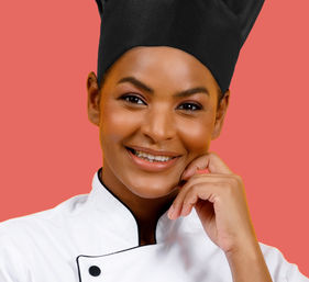 Smiling chef in a black toque and white double-breasted jacket, hand resting on chin in a close-up studio portrait on a coral background
