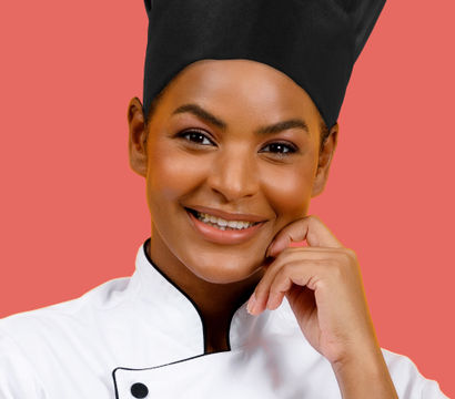 Smiling chef in a black toque and white double-breasted jacket, hand resting on chin in a close-up studio portrait on a coral background