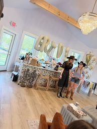 Two women in cowboy hats and boots stand in a bright, country‑chic event space with gold "BRIDE" balloons, hat wall, gift table and crystal chandelier over wood floors and a tiled bar counter.