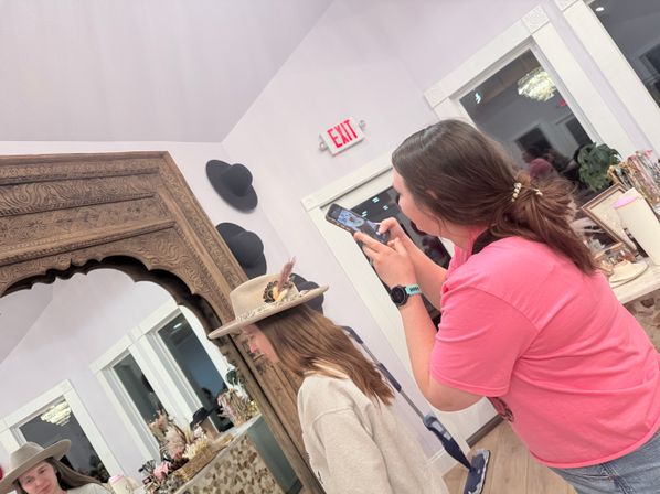 In a boutique hat shop, a person in a pink shirt photographs a companion trying on a feather-trimmed felt hat in front of an ornate wooden mirror and hat display.
