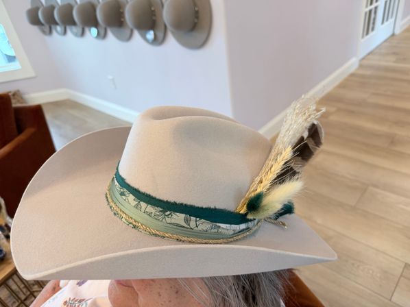 Stylish cream felt cowboy hat with a green patterned hatband, braided cord and feather-and-wheat stalk accent, shown indoors on a head against wood floors and a wall of hat hooks.