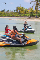 Three women in bright swimsuits riding colorful jet skis across a calm tropical lagoon near a palm-lined sandy beach — fun watersports vacation scene.