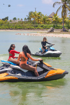Three women in bright swimsuits riding colorful jet skis across a calm tropical lagoon near a palm-lined sandy beach — fun watersports vacation scene.