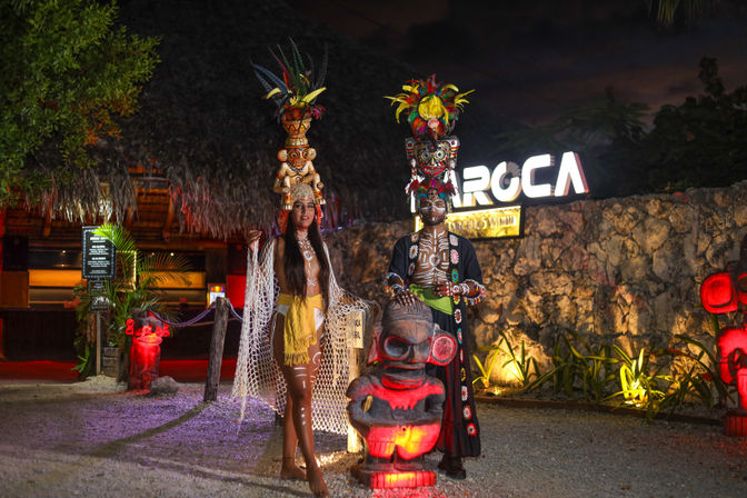 Vibrant nighttime scene of two performers in tall feathered headdresses and tribal body paint posing with illuminated carved tiki statues at a thatched-roof tropical beach venue entrance.