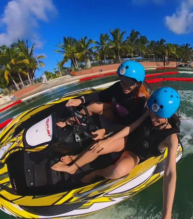 Two people wearing blue helmets riding a yellow jet ski in a sunny tropical lagoon with palm trees and bright blue sky