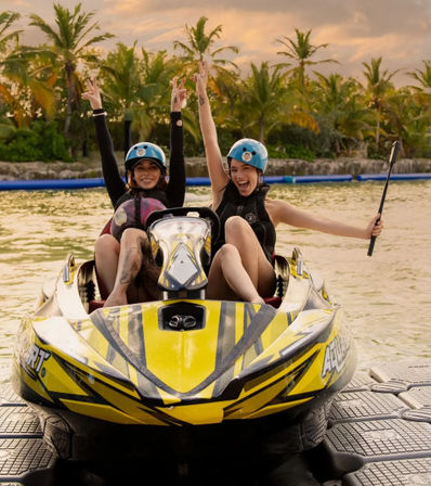 Two friends wearing helmets cheering on a yellow two-person jet ski at a tropical lagoon with palm trees and sunset, docked on a floating platform — water sports fun