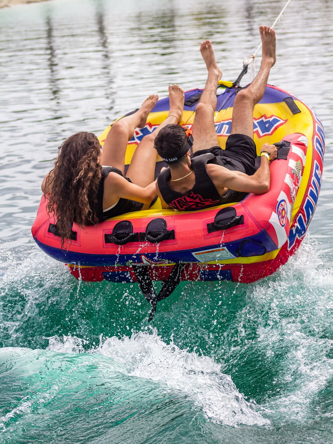 Two people in life jackets riding a colorful towable tube over a boat wake on a turquoise lake, splashing water — summer lake tubing fun