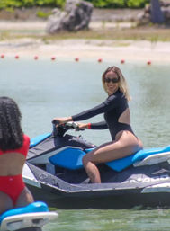 Smiling woman in a black swimsuit and sunglasses riding a blue jet ski on clear coastal water near a sandy beach with buoy markers — upbeat summer watersport scene.