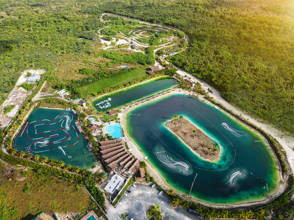 Aerial view of a tropical waterpark with turquoise man-made lagoons, a sandy central island, palm-lined shores, winding jet-ski wake trails and obstacle course, set against dense green forest.