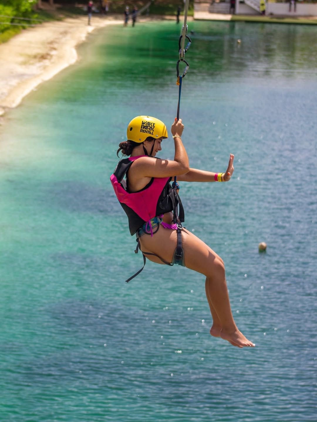Zipliner in a yellow helmet and pink life vest suspended in a harness above a turquoise lake near a sandy shore — outdoor water adventure