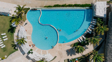 Aerial view of a curvy turquoise resort pool at a tropical hotel, framed by palm trees, rows of white lounge chairs and people relaxing on colorful inflatables.