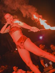 Fire dancer in red costume and tribal face paint twirling flaming torches in an outdoor nighttime performance with smoke and a watching crowd — live fire show entertainment.