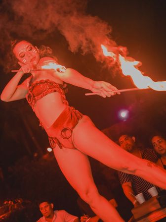 Fire dancer in red costume and tribal face paint twirling flaming torches in an outdoor nighttime performance with smoke and a watching crowd — live fire show entertainment.