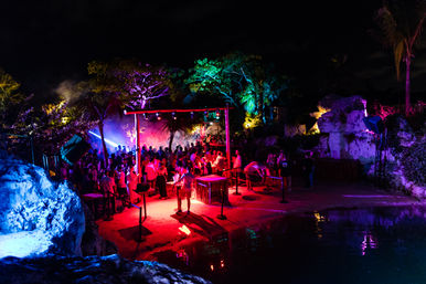 Nighttime tropical beach party at a lagoon with a crowded dance area, neon red-blue-green lighting, palm trees, DJ setup and colorful reflections on the water