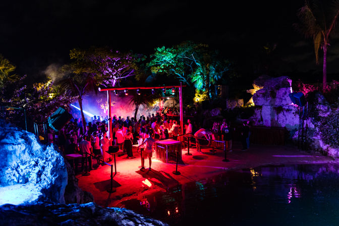 Nighttime tropical beach party at a lagoon with a crowded dance area, neon red-blue-green lighting, palm trees, DJ setup and colorful reflections on the water